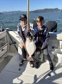 Two young anglers proudly displaying a large Halibut on a boat during a fishing trip.