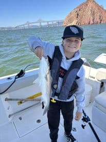 A young angler proudly displaying a striped bass while fishing on a boat near a scenic bridge.
