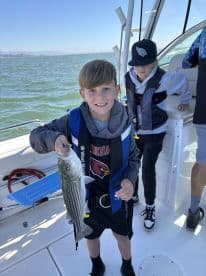 A young angler proudly displaying a striped bass while fishing on a boat, surrounded by fellow young anglers.