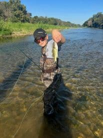 A young angler proudly displaying a freshly caught Largemouth Bass while standing in a clear river, showcasing a beautiful day of fishing.
