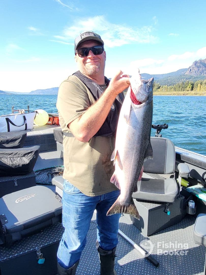 An angler proudly displaying a large Salmon while fishing on a scenic lake, surrounded by lush greenery and mountains.
