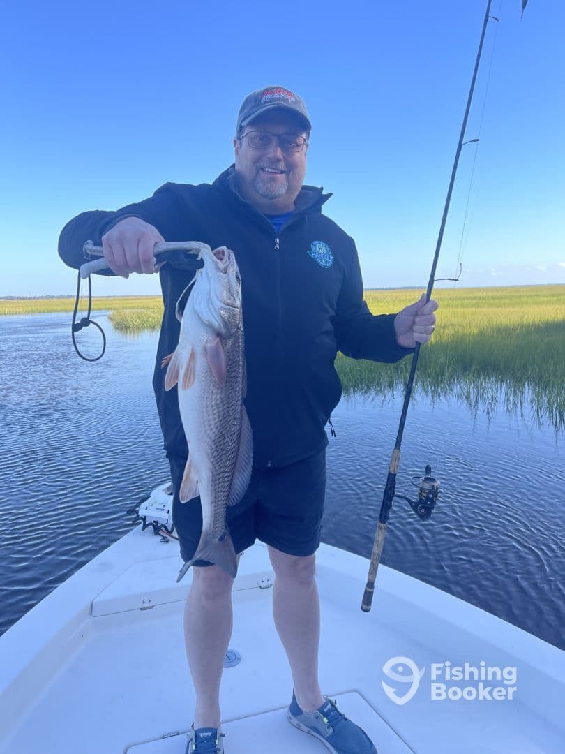 An angler proudly displaying a large Redfish while fishing from a boat in a scenic coastal environment.