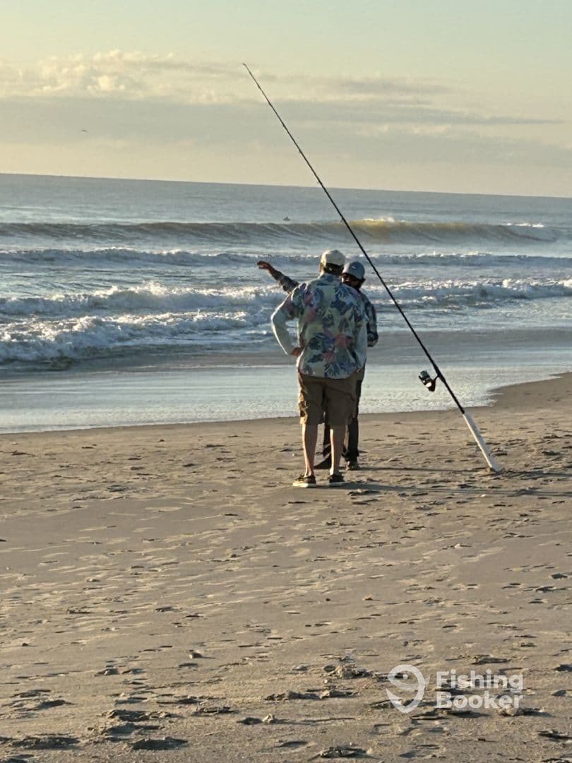 Two anglers discussing fishing techniques while surf fishing on a sandy beach during sunset.