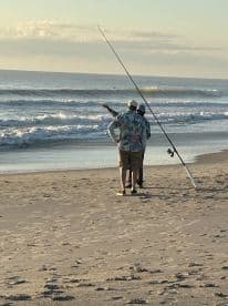 Two anglers discussing fishing techniques while surf fishing on a sandy beach during sunset.