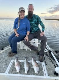Two anglers sitting on a boat with three freshly caught Redfish displayed in front of them, enjoying a serene fishing experience.