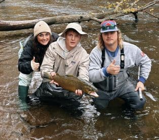 Three anglers celebrating a successful catch of a large Brown Trout while wading in a river, showcasing teamwork and excitement.
