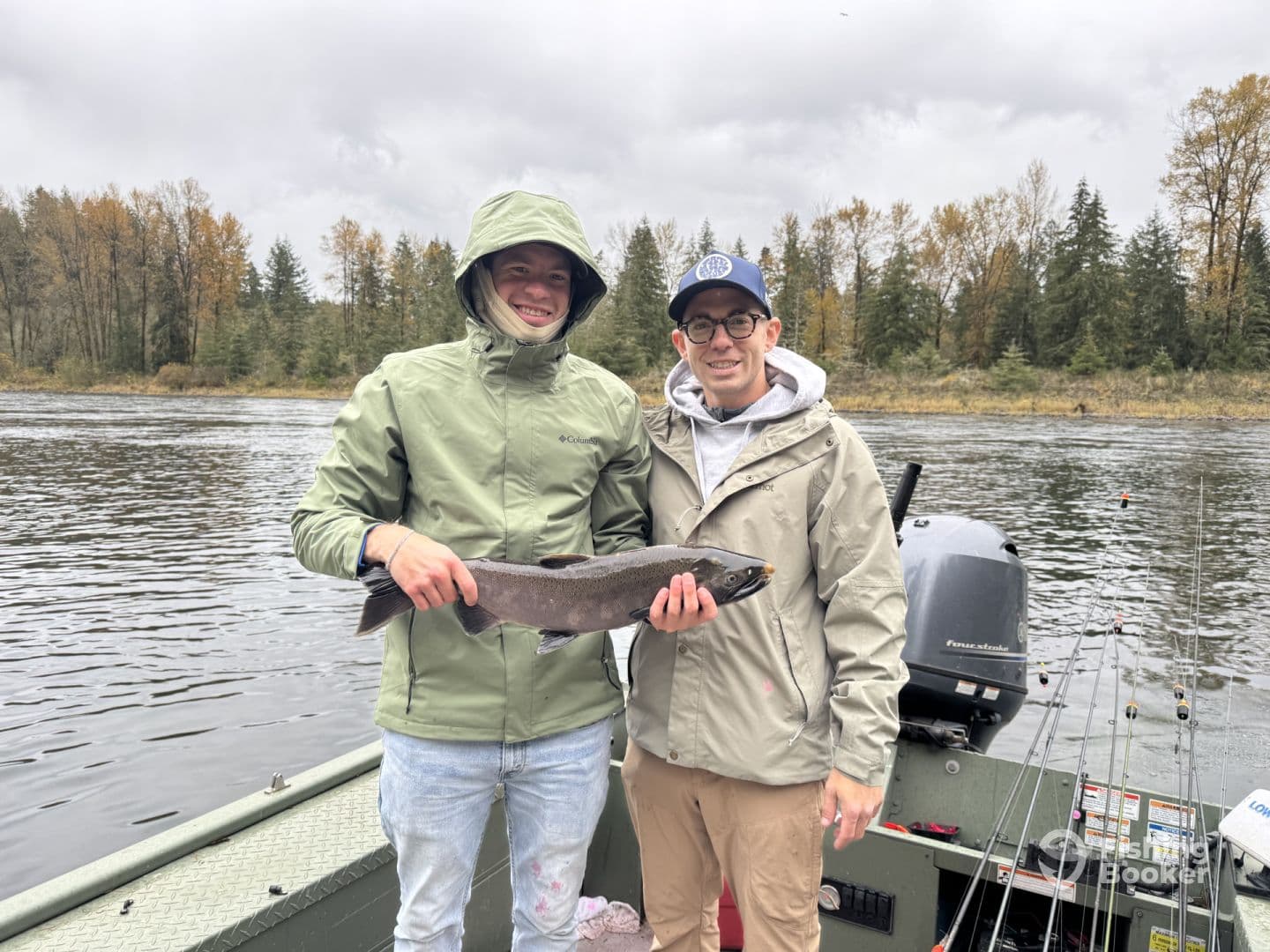 Two anglers proudly displaying a freshly caught Steelhead while fishing on a river, surrounded by a scenic forest backdrop.