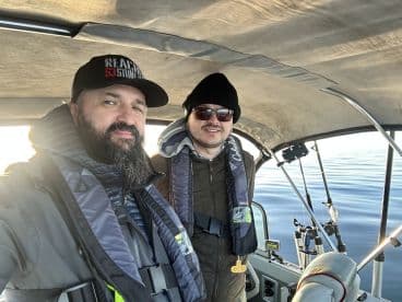 Two anglers inside a fishing boat, equipped with life jackets, enjoying a calm day on the water.