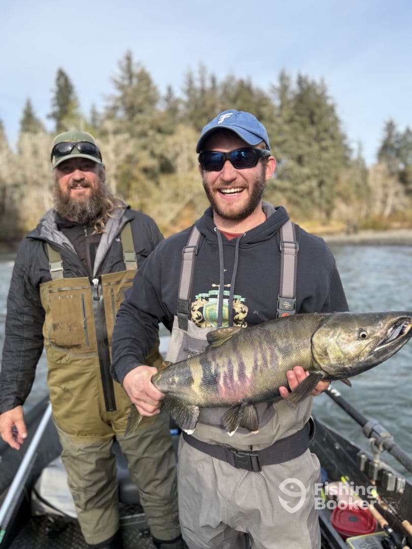 Two anglers proudly displaying a large Chinook Salmon while fishing in a scenic river environment.