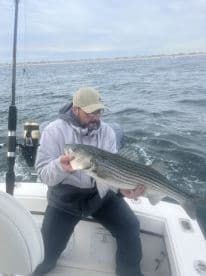 An angler proudly displaying a large Striped Bass while fishing on a boat, with a coastal backdrop.