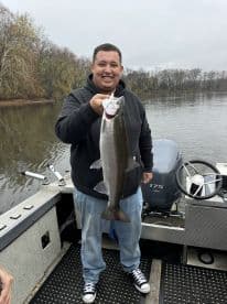 Angler proudly displaying a large Steelhead trout while fishing from a boat on a calm river.