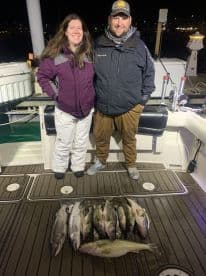 Two anglers proudly displaying their catch of various fish on the deck of a fishing boat at night.