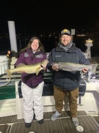 Two anglers proudly displaying their Walleye catches on a boat at night, showcasing a successful fishing trip.