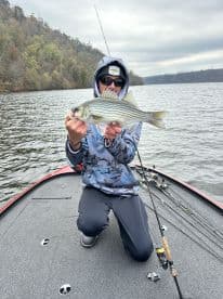 A young angler proudly displaying a striped bass while kneeling on a fishing boat in a serene lake setting.