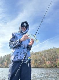 Angler holding a freshly caught Crappie while fishing on a serene lake surrounded by autumn foliage.
