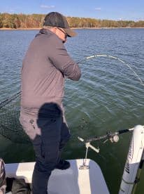 Angler reeling in a fish while fishing from a boat on a calm lake, surrounded by autumn foliage.