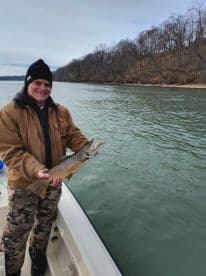 A proud angler holding a Brown Trout while fishing from a boat on a calm river, showcasing a successful catch.