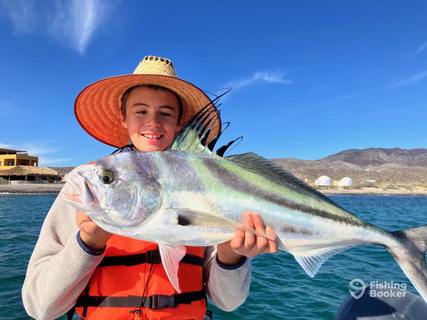 A young angler proudly displaying a large Roosterfish while fishing from a boat in a scenic coastal environment.