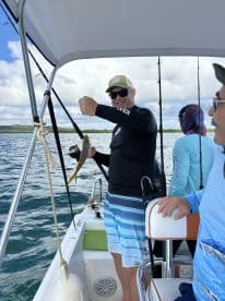 An angler joyfully reeling in a fish while fishing on a boat, surrounded by fellow anglers in a scenic waterway.
