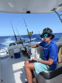 A young angler enjoying snacks while seated inside a fishing boat, surrounded by fishing rods and the open ocean in the background.