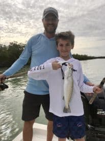 A young boy proudly displaying a Speckled Trout while fishing with a family member on a boat in a scenic coastal environment.