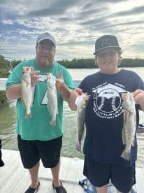 A proud father and son duo displaying their catch of Speckled Trout at a scenic dock, showcasing a successful fishing trip.