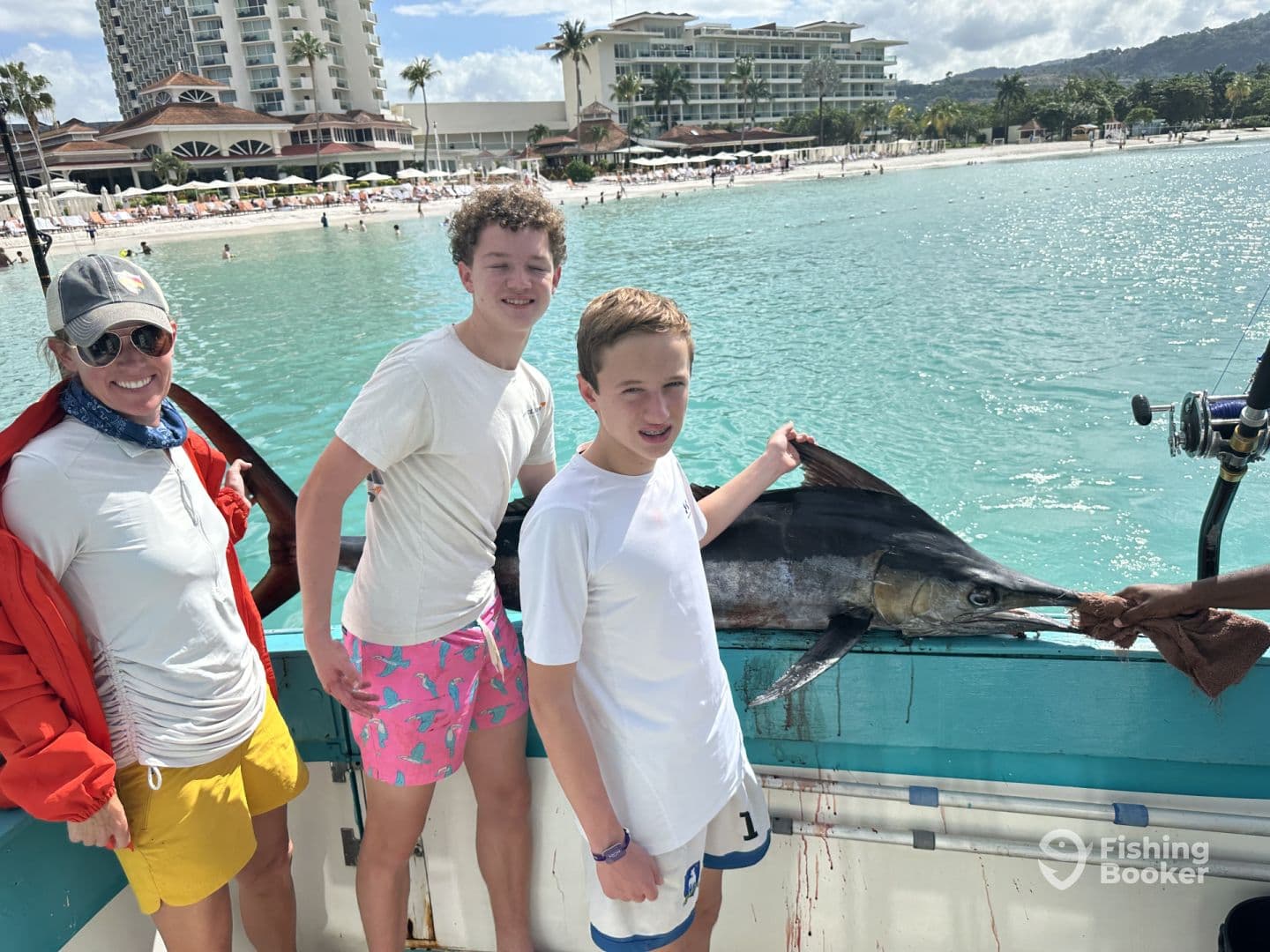 A family proudly displays a large Marlin on a fishing charter in a tropical setting, with a beautiful beach and resort in the background.