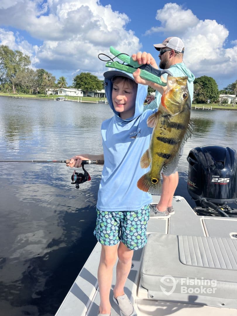 A young angler proudly displaying a colorful Peacock Bass while fishing from a boat in a scenic freshwater environment.