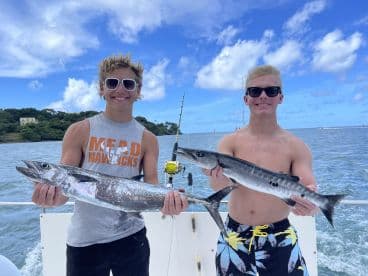 Two young anglers proudly displaying their catches of Barracuda while fishing on a boat in clear blue waters.