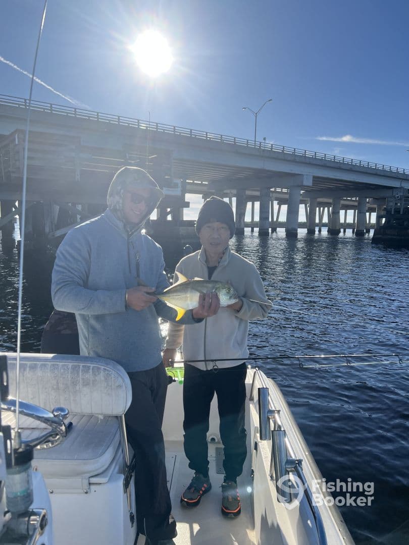 Two anglers proudly displaying a caught fish while on a boat under a bridge, enjoying a sunny day on the water.