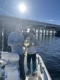Two anglers proudly displaying a caught fish while on a boat under a bridge, enjoying a sunny day on the water.