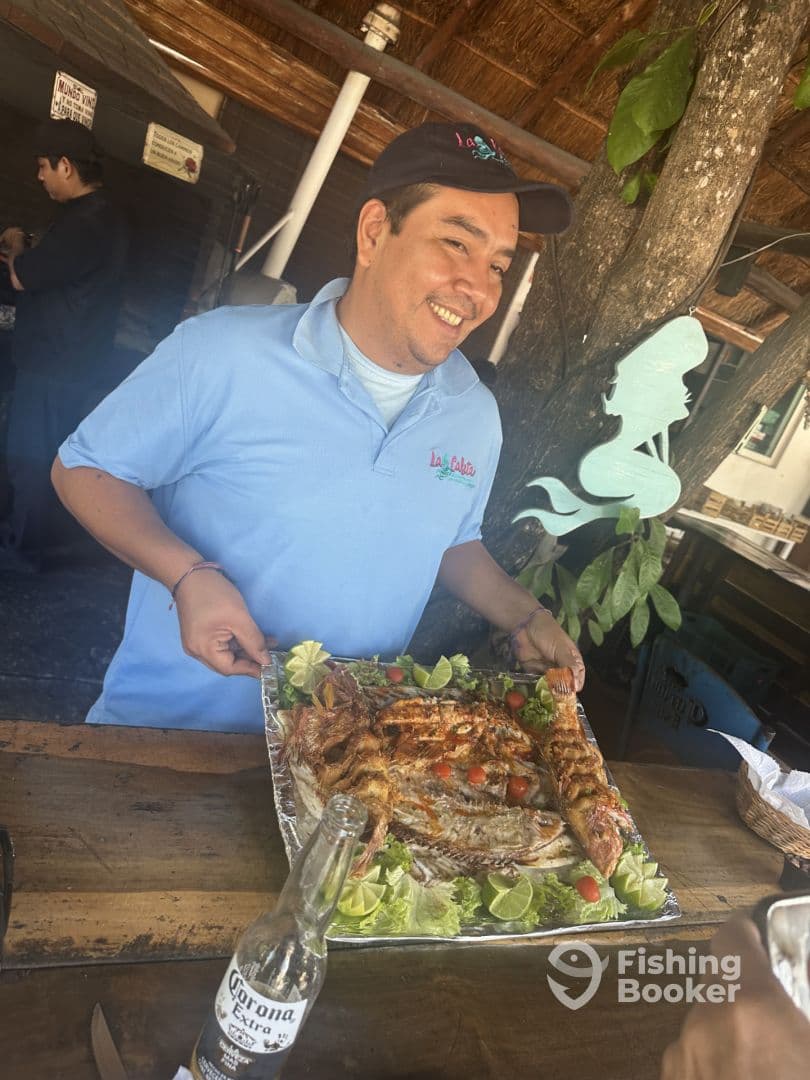 A smiling individual holding a platter of grilled fish garnished with lime and vegetables, showcasing a culinary experience at a fishing-related establishment.