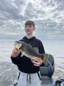 A young angler proudly displaying a large Largemouth Bass while fishing on a calm lake, showcasing a beautiful cloudy sky.