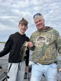 A young angler and an adult proudly displaying a large Largemouth Bass while fishing on a calm waterway.