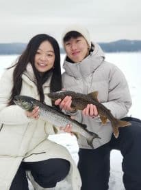 Two young anglers proudly displaying their catches of trout while ice fishing on a snowy lake.