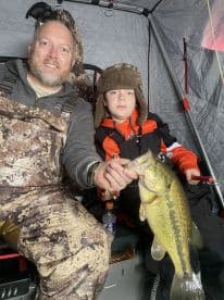 A young angler and an adult proudly displaying a large Largemouth Bass while ice fishing in a cozy shelter.