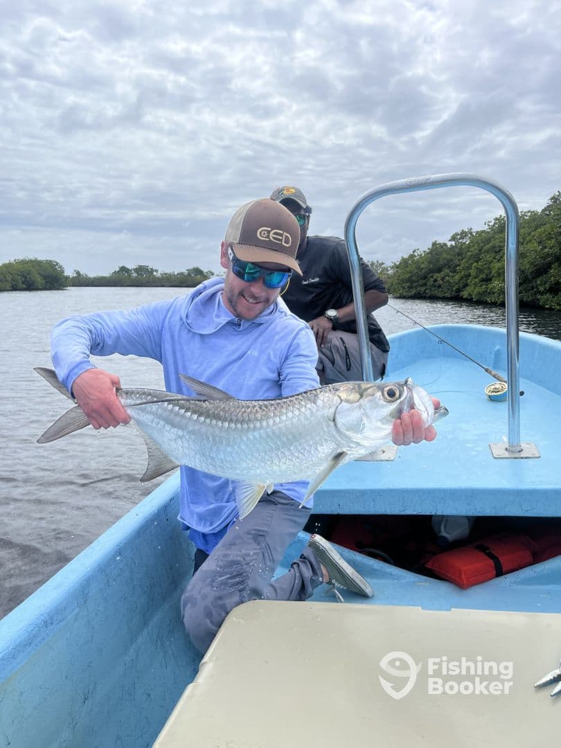 Angler proudly displaying a large Tarpon while fishing from a small boat in a mangrove-lined waterway.