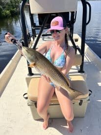 A smiling angler proudly displays a large Redfish while seated on a boat, enjoying a sunny day on the water.