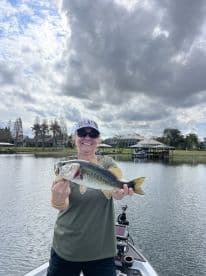 An angler proudly displaying a large Largemouth Bass while fishing from a boat in a scenic lake setting.