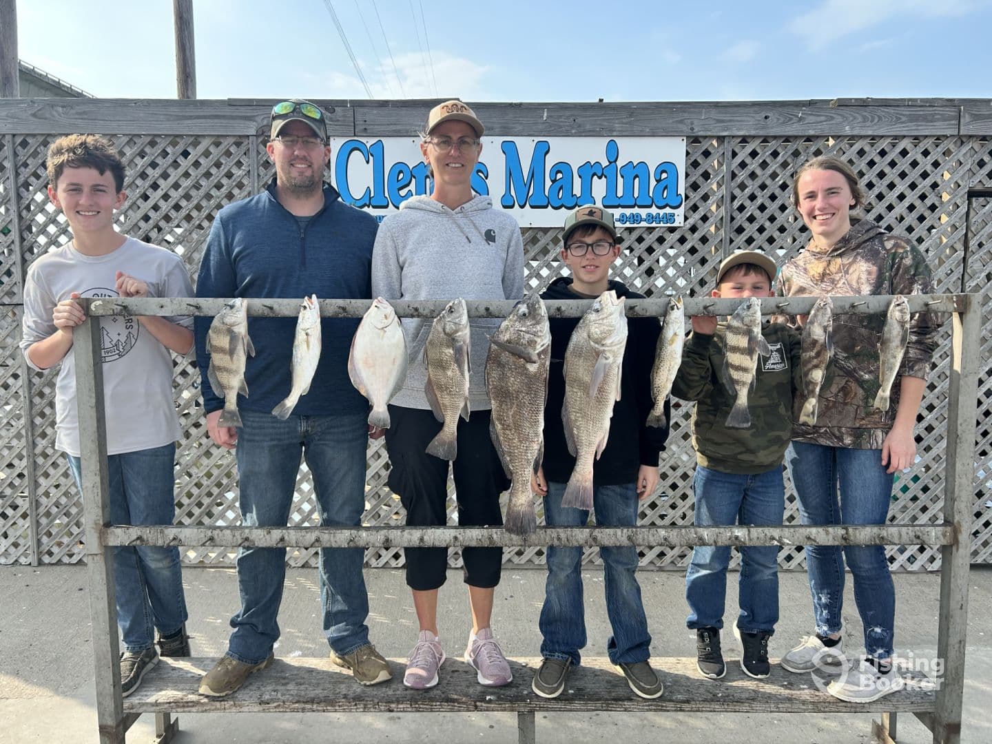 A family poses proudly in front of a catch board displaying various fish, including flounder and trout, at a marina after a successful fishing trip.