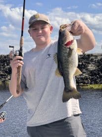 A young angler proudly displaying a large Largemouth Bass while fishing in a scenic freshwater environment.