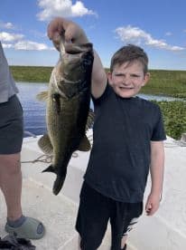 A young angler proudly displaying a large Largemouth Bass while fishing on a scenic waterway.