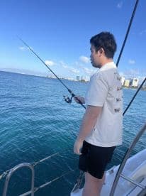 A young angler casting a line from a boat in clear blue waters, with a city skyline in the background.