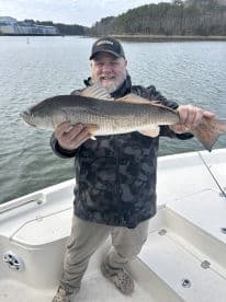 An angler proudly displaying a large Redfish while on a fishing trip, with a scenic backdrop of a calm waterway.