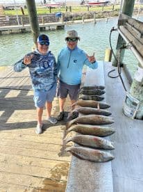 Two anglers proudly displaying their catch of several Redfish on a cleaning table at a coastal fishing dock.