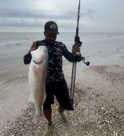 Young angler proudly holding a large Redfish on the beach, showcasing a successful catch during a coastal fishing trip.