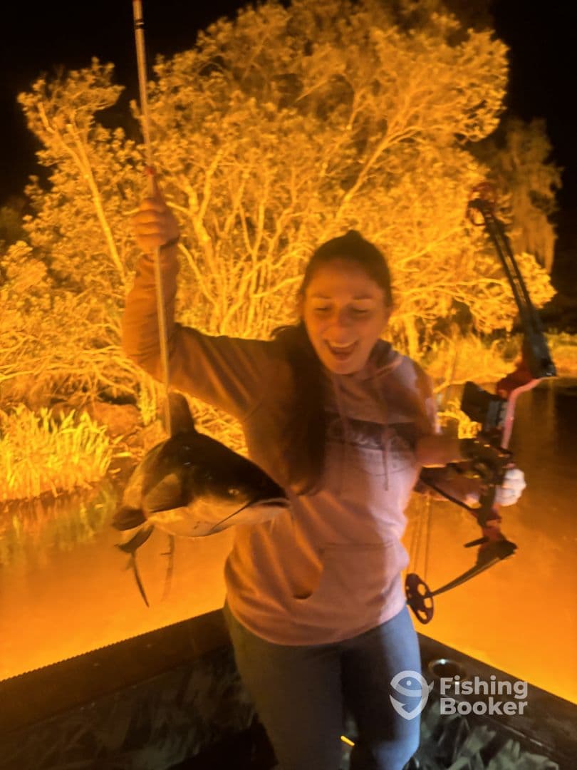 Angler joyfully displaying a large catfish while bowfishing at night, surrounded by a vibrant natural backdrop.