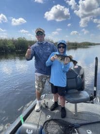 A young angler proudly displaying a large Largemouth Bass while fishing on a boat in a scenic freshwater environment.