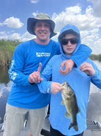 A young angler proudly displaying a large Largemouth Bass while fishing with an adult companion on a calm waterway.