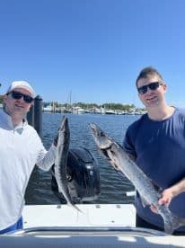Two anglers proudly displaying their catches of Barracuda while on a fishing trip, with a marina backdrop.